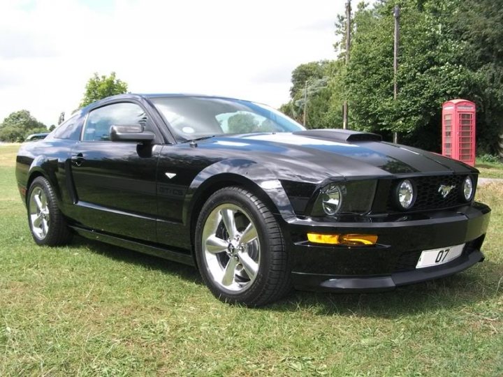 Importing to UK - Page 1 - Mustangs - PistonHeads - The image shows a black Mustang car parked on a green grass field. The car is facing towards the left side of the frame, capturing the front portion of the vehicle. It appears to be a convertible model, distinguished by its black roof that is currently up. The license plate of the car reads "09 07", and a small detail unique to convertible Mustangs is a solar panel-like accessory on the trunk. In the background, partially obscured by the car, there's a red telephone booth. The overall setting suggests an outdoor, perhaps recreational area.