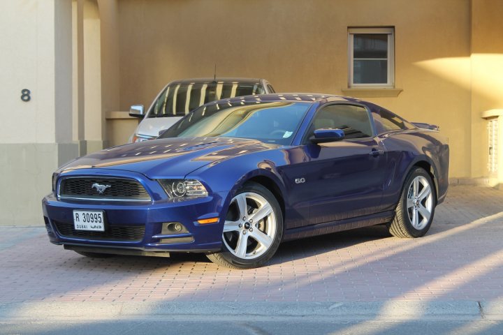 Sold the Golf, replaced with proper car - Page 1 - Middle East - PistonHeads - The image captures a scene in a parking lot, where a striking blue Mustang and a white SUV are parked next to a beige building. The Mustang is stationary, facing to the driver's left, with the white SUV positioned behind it. The building's facade serves as a backdrop, with a window that suggests it might house a vehicle sales or service area. The license plates of both vehicles are clearly visible, adding authenticity to the scene.