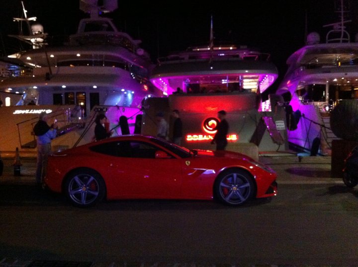 Pistonheads - In the center of the dock, a vibrant red Ferrari sports car is parked, its glossy finish reflecting the ambient light. To the left, two large boats are docked, their silhouettes towering over the Ferrari. An array of people can be seen walking around the boats, adding a sense of activity to the scene. The ground underneath is a dark shadow, considered it's night time. In the background, a car in motion can be seen, suggesting the hustle and bustle outside this tranquil dock scene.