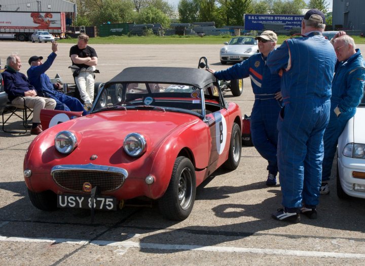 Seeking owners... - Page 1 - London - PistonHeads - The image depicts a gathering of people around a vintage red sports car at a parking lot. The car, bearing the license plate "USSL 875," attracts the attention of the group, who are dressed in matching uniforms. The vehicle is positioned in the middle of the image, with some individuals conversing while others are inspecting its features. The setting appears to be an outdoor event, with cars and equipment in the background, possibly suggesting a car show or motorsport venue.