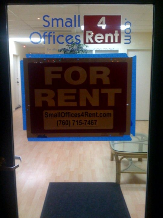 Rent Sign - This image reveals a sign in a door window that advertises a 'For Rent' property, located at '150 (760) - 75467'. The sign also gives the phone number 'SmallOffices4Rent.com', suggesting that interested parties can call or visit this website for more information or inquiries related to the rental property. The door itself is made of glass, providing a clear view of the interior, where a lamp and furniture can be seen. The sign is covered with a piece of blue tape, possibly indicating unavailability or a construction or repair process ongoing inside the rented property.