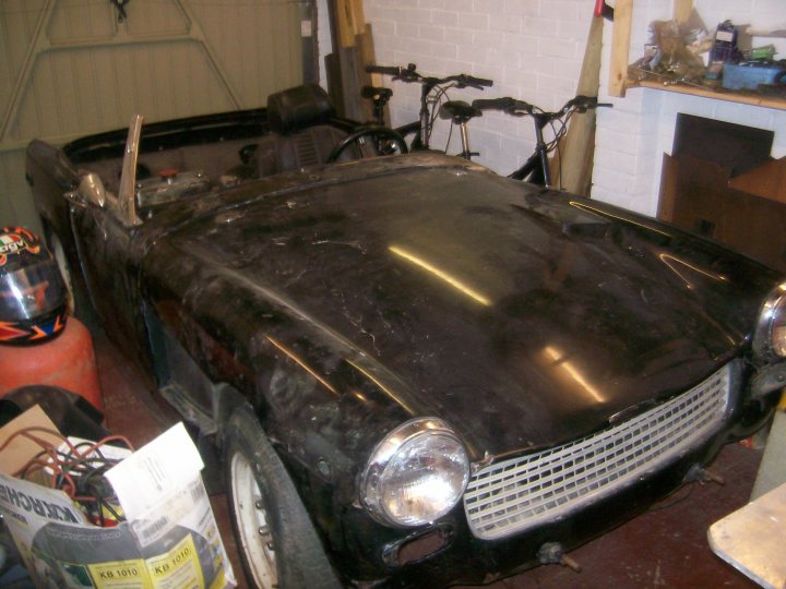 Austin Sprite Healey Pistonheads - The image showcases a vintage car, covered in a black canvas and displayed indoors. The interior of the car has been removed, revealing a skeletal structure of unpainted metal beneath. A bicycle is positioned to the left of the car, while the rear of another bike is visible on the right. The car is parked next to a white wall, adding a contrasting background to the vintage vehicle. The setting suggests a workshop or a garage dedicated to the maintenance or restoration of vehicles.