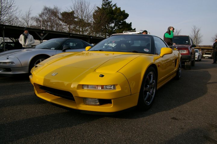 Goodwood Breakfast Club - Sushi Sunday - photos - Page 1 - Goodwood Events - PistonHeads - The image shows a gathering of cars on a tarmac, with a bright yellow Honda S2000 sports car prominently featured in the foreground. A man is standing nearby, gesturing towards the car. In the background, several other people and cars can be seen. The scene suggests a car meet or exhibition event, where car enthusiasts can display and crowd around vehicles of interest. The focus is on the cars, especially the vivid yellow sports car that stands out among the other vehicles. The atmosphere is likely that of a car show or meetup, with attendees enjoying the vehicles on display.