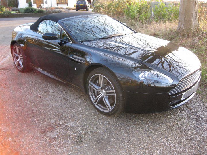 Back in the fold and its feeling good!!!! - Page 1 - Aston Martin - PistonHeads - In the image, a sleek black sports car is parked on an asphalt driveway. The car's retractable hard top roof is retracted, revealing the interior. The car's rims are visible, and it appears to be parked near a grassy area and under what might be sunlight. There are some blurred objects and scenery reflected in the car's surface, suggesting a landscaped environment around it.