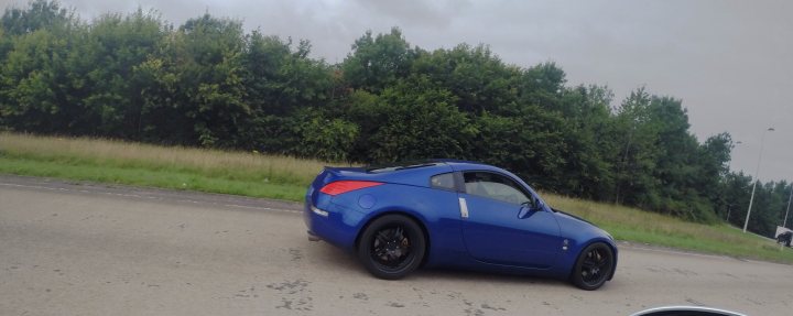 Meet, Drive, Photos and Drone 16th August 2015 - Page 9 - South Wales - PistonHeads - The image depicts a bright blue sports car driving on a road with a grassy area to the left. The sky overhead appears to be cloudy, and the time of the day might suggest it's either early morning or late evening, given the soft, diffused lighting. Several tall green trees line the road, providing a natural backdrop to the scene. The image is slightly fuzzy, which could indicate motion blur from the high-speed movement of the car.