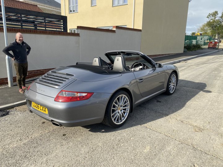 In the image, a silver sports car is parked on a road beside a residential building. The car appears to be in good condition with its top down, revealing an interior that seems ready for a drive. The driver's side door is open, suggesting that someone might have just exited or is about to enter. The setting is calm and quiet, with no other vehicles visible in the immediate vicinity. A single individual can be seen standing near the car, possibly associated with it. The overall scene suggests a peaceful suburban environment where the car is parked temporarily.