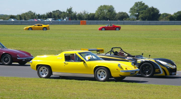 Snetterton Lotus Festival 25/26 June - Page 1 - General Lotus Stuff - PistonHeads - The image shows a vibrant scene of car racing on a grass track. There are four distinct vehicles in total, each showcasing a different design and color. Three of the cars have yellow and black designs, possibly representing racing teams, while a single red car brings a splash of contrasting color to the scene. The track is winding through a rural landscape, with trees and grassy areas visible in the background. The bright daylight suggests it's a fair-weather racing event. Each car has its roof down, allowing the drivers an unrestricted view of the track ahead. The stance of the cars suggests they're in motion, adding dynamism to the scene.