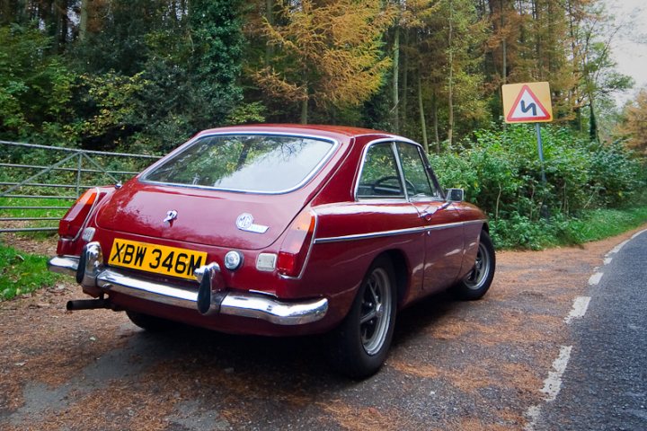 A red car is parked in a parking lot - Pistonheads - This image portrays a vibrant red classic car parked on the side of a road in a rural setting. The vehicle appears to be a MGA, a classic two-door sports car, identifiable by its distinctive rounded shape, chrome bumpers, and black side curtains. The car is parked on a gravel shoulder, which has a signpost with a black and red warning sign, depicting a car with arrows pointing right. Behind the car, the scene is filled with lush greenery, fallen leaves, and trees, suggesting it might be autumn. The overall ambiance of the image suggests a peaceful day in the countryside.