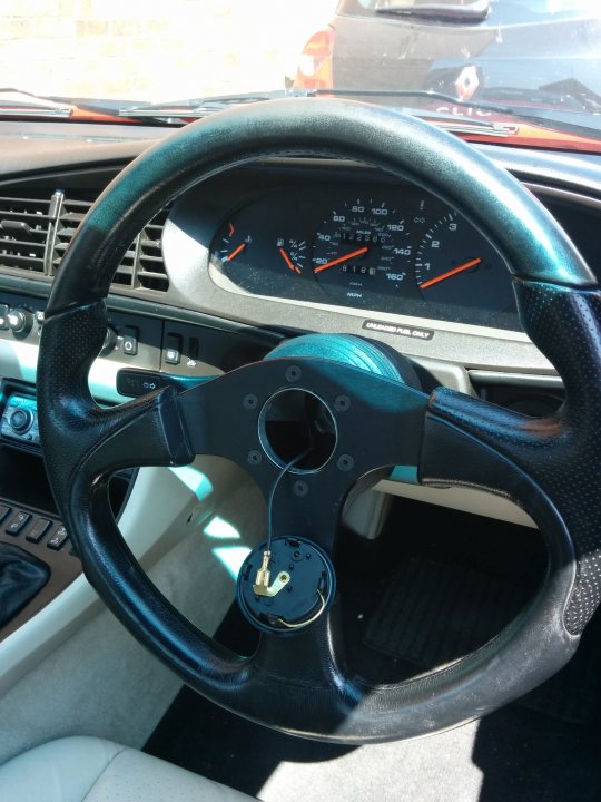 Momo steering wheel - Page 1 - Front Engined Porsches - PistonHeads - The image displays the inside of a car's cockpit, focusing on the steering wheel. The steering wheel is black with a silver center and appears to have a logo, possibly suggesting brand affiliation. Behind the wheel, there is a dashboard with a display showing "80" at the top, which could indicate the speed of the car or another relevant setting. To the left of the wheel, there is a small section of the car's dash visible with what seems to be the gear shift lever and other instrumentation. The viewer's perspective implies that they are either seated in the passenger seat or standing in the doorway of the car, looking into the cockpit.