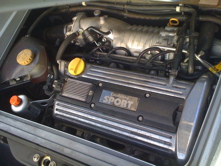 Pistonheads Courtenays Converted - The image is taken from a perspective looking down on a engine compartment. It shows an open hood revealing various mechanical parts. Prominently placed on the engine are a yellow button and a water cap. There is a close-up view of a metal component with text that reads "PORT ECU," indicating that this is a part of the engine control unit. The precise location of the components and the specific actions they perform are not clear from the image alone.