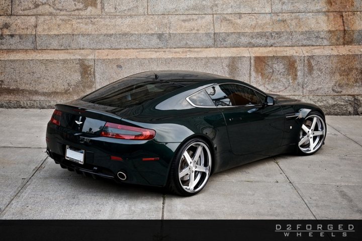 Car Porn - Page 1 - Aston Martin - PistonHeads - The image showcases a sleek, black sports car parked on a concrete pavement sloped against a backdrop of a concrete building. The car's design is accentuated by a visible rear spoiler and a set of multispoke wheels. The vehicle's license plate is clearly visible. Above the car, the text "D2FORGED WHEELS" suggests a partnership or sponsorship.