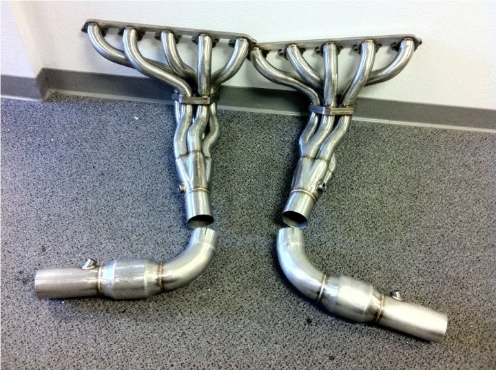 Exhaust Query Cat Pistonheads - The image shows two pipes resting on a gray carpeted floor, positioned against a white wall. The pipes are long and metallic, and they are carefully arranged to look like sculptural pieces, as if they are intertwining together or forming an abstract design. The pipes appear well-cleaned and polished, suggesting attention to detail. This setup gives an impression of art or design elements in an otherwise ordinary room.