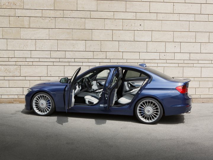 RE: Alpina B3 Bi-Turbo: Review - Page 2 - General Gassing - PistonHeads - The image displays a dark blue, four-door sedan with the rear door open, parked next to a light-colored brick wall. The car is positioned in front of a building with an architectural appearance. The visible interior through the open door suggests leather upholstery and chrome accents. The pavement beneath the car is gray, and the lighting appears to be overcast, creating soft illumination on the vehicle.