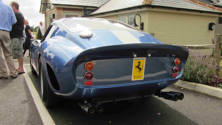 Hare Breakfast Meet - August - Page 1 - Kent & Essex - PistonHeads - The image features a vibrant blue classic car with a white stripe down the middle of its hood. This striking vehicle is parked on the side of the road, adjacent to the curb. There are two visible individuals in the background, suggesting a public or communal setting. The weather appears to be in the range of cloudy, and the presence of a house in the background adds a pinpoint to the location.