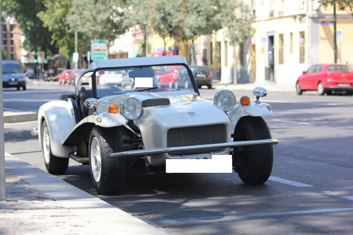 Lotus 7 - S4 - Page 27 - Caterham - PistonHeads - The image depicts an old-fashioned white classic car, possibly a Bugatti, parked on a busy street. The car's vintage design, featuring a convertible top and exposed chrome detailing, stands out against the modern backdrop of the city. The city street itself is lining both sides with stoops and automobiles parked on the curb. The traffic suggests it's a bustling area, with pedestrians and vehicles interacting and moving around each other, creating a lively urban scene.