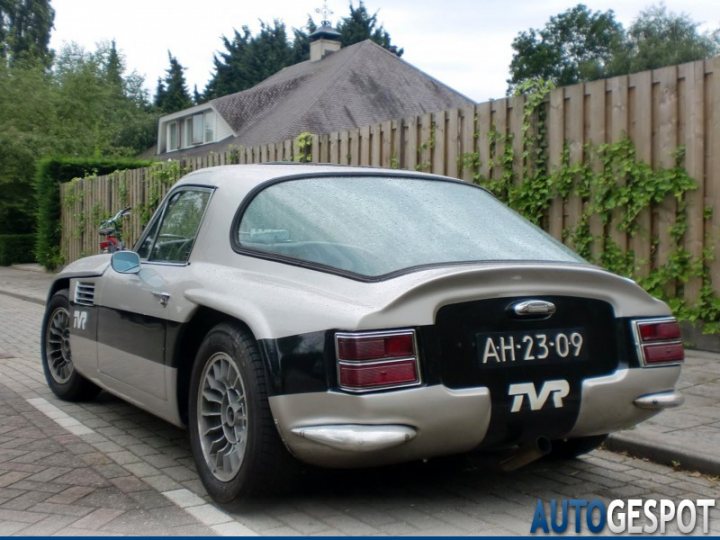 Early TVR Pictures - Page 81 - Classics - PistonHeads - The image showcases a sleek sports car parked on a driveway adjacent to a wooden fence. The car has a metallic finish, divided between a light and dark gray hue, with a black and white stripe running along the side. The rear light bar is a prominent feature, giving the car a distinctive look. The license plate reads "TV R", indicating the brand or model of the car. The setting seems peaceful, with greenery visible in the background, suggesting a well-maintained garden or residential space.