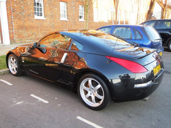 350/370z - Page 1 - Jap Chat - PistonHeads - This image shows a dark blue car parked on an asphalt surface next to a sidewalk. Its design has a stylish black finish on the hood and roof, and the interior is revealed through the windows, featuring a black interior as well. The background of the image includes a steeple, indicating that the car is parked in front of a building with traditional architecture, set against a clear sky.