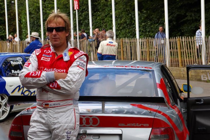 FOS good photography vantage points.. - Page 1 - Goodwood Events - PistonHeads - The image features a man standing beside a silver and red Audi R8 sports car. He is wearing a white driver's uniform with red and white initials, sunglasses, and appears to be in confidence or assertiveness as he crosses his arms. The man has short blonde hair. In the background, there is a blur of other individuals, suggesting a public setting. The atmosphere seems lively and crowded, likely indicating an event or public gathering. There are also small red and white banners in the distance.