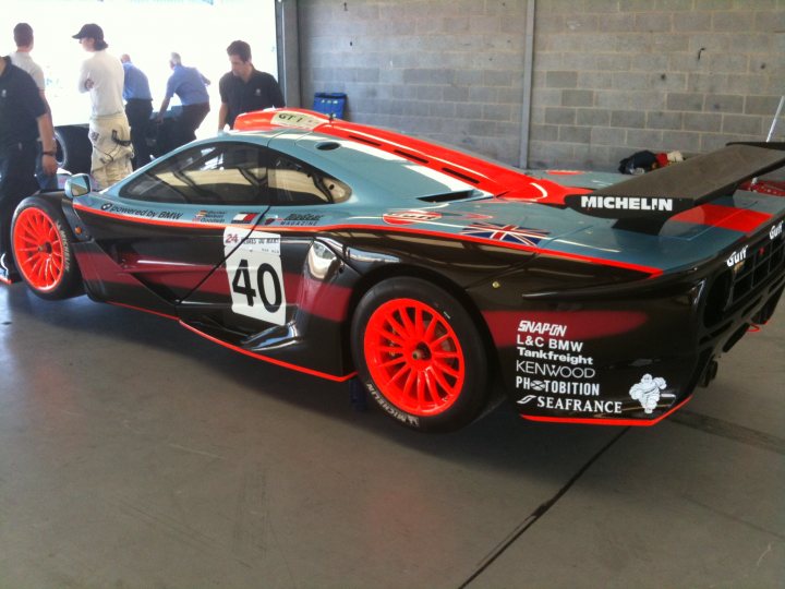 Mclaren Flemke Pistonheads - This image features a race car, predominantly black and red with orange rims, parked in a garage or workshop setting. The car has the number "40" prominently displayed, and there are various text advertisements along the side. There are five people visible in the background, three of which appear to be workers, standing by the car and the garage, possibly admiring or discussing the vehicle. The car shows signs of use and wear, suggesting it has seen action on the race track. The overall image conveys the environment of a motorsport team's garage, with the car being the central focus.