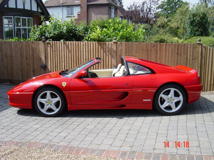 Had a fezza wobble - Page 1 - Aston Martin - PistonHeads - This image portrays a striking red Ferrari convertible parked in a driveway. The car's top is down, revealing its interior. The Ferrari features a sleek design with alloy wheels and is positioned against a vertical wooden slat fence. The car's vibrant color contrasts with the more muted tones of the brick driveway it sits on. In the background, there are greenery and a building, adding context to the scene.