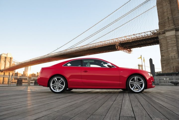 Wolves Pistonheads Clothing Sheep - The image captures a striking red sports car parked on a wooden pier, facing towards the right. The car is positioned in front of the iconic Brooklyn Bridge, with the bridge tower stretching into the sky in the background. The wooden pier offers a contrasting texture to the sleek form of the sports car. The overall mood of the image is one of calmness and urban beauty.