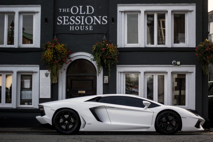 The GT Cafe - the ace cafe of the north - Oct Meet - Page 1 - North West - PistonHeads - The image showcases a white Lamborghini sports car prominently parked outside a black building. The car is positioned on the right side of the frame, its sleek design contrasting with the building's vertical lines and multiple windows. Above the car, the building walls are adorned with hanging plants and a sign with the text "The Old Sessions House" indicating the building's purpose. The scene exudes an urban, possibly European, ambiance.