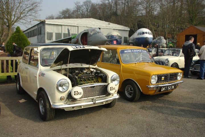 Day Brooklands Pistonheads Mini - This image captures a scene of two iconic cars, the Mini Cooper and the original Volkswagen Beetle, parked side by side. The Mini Cooper, painted white with its hood up, is on the left. It's closer to the camera, making it more prominent in the image. The white Volkswagen Beetle, standing out against its yellow background, is on the right. Both vehicles are parked on a concrete surface, with the building and trees providing a backdrop to the vintage automobiles. A few individuals can be seen in the background, indicating a public setting or event where these classic cars are on display.