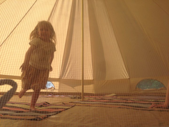 Bell tent ..... Hold my hand please - Page 1 - Tents, Caravans & Motorhomes - PistonHeads - The image captures a young child standing under a large white tent. The child is dressed in light-colored clothing and appears to be smiling. The tent is erected on a flat surface with a patterned carpet underneath, providing a vibrant contrast to the white fabric of the tent. To the right, there's a glimpse of a second person, though only their arm and part of their head are visible. The overall scene is one of relaxation and enjoyment, likely a festival or outdoor event setting.