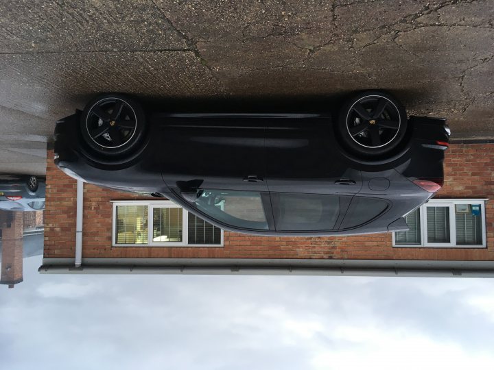 Cayenne 4.2 Diesel - Page 1 - Porsche General - PistonHeads - The image shows a dark-colored sports car parked at an angle. It features a shiny exterior and black rims on the wheels. The vehicle is positioned in front of a building with a brick facade, indicating it might be an urban or residential setting. The car's perspective is slightly distorted due to its position near a mirror or similar reflective surface, which gives the image a unique, almost mirrored appearance.