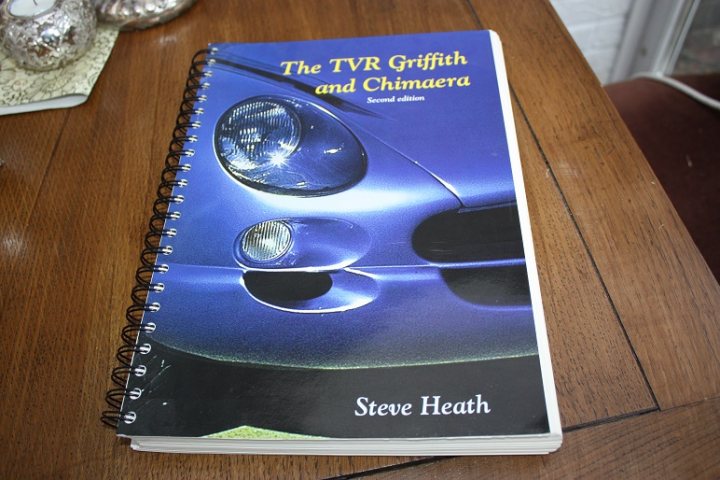 NE TVR Chimaera or Griff owners? - Page 1 - North East - PistonHeads - The image displays a book with a cover that features a photograph of a car's front left side, specifically the headlight and grille. The title of the book, "THE TVR GRIFFITH AND CHIMAERA," is written in large text at the top of the cover. Below the title, the subtitle "Second Edition" is indicated. The author's name, Steve Heath, appears at the bottom. The book is resting on a wooden surface, and there are other items in the background, such as what appears to be a silver jewelry box and a piece of paper with a patterned design. The style of the photograph is straightforward, and the focus is on the book and its cover.