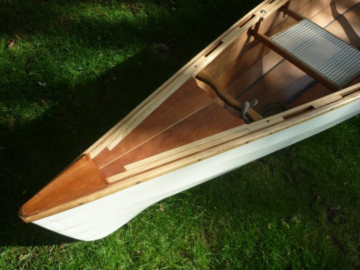 A red and white boat sitting on top of a lush green field - The image shows a small wooden boat resting on grass. It features a pointed bow and a flat stern, with a prominent seat in the center. The boat's structure reveals layers of plywood panels, creating an intricate visible weave. There is no one around or in the boat. The background consists mainly of green grass, suggesting an outdoor setting.