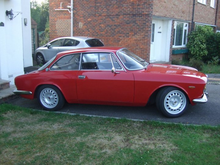 Lets  Alfas Pistonheads - The image depicts a vibrant red classic car parked on the grass outside a building. The car, which appears to be a model Ford Cortina, is characterized by its silver-colored roof. The surrounding area includes a brick wall and a pathway leading to a white door in the distance. There's a door with a window next to the parked car, suggesting this photo might have been taken at a house or an apartment building. Another car is parked further down the driveway, and a lush patch of verdant grass provides a serene backdrop. The overall scene conveys a sense of nostalgia, as classic cars like this one are often associated with the past.