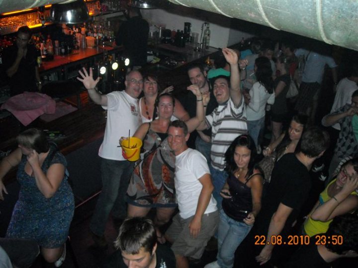 Pistonheads - The image shows a lively indoor scene, likely in a bar or club, where a large group of people are gathered on a dance floor. The atmosphere appears to be jovial, with individuals posing and celebrating. Centered in the image, a diverse group of six individuals is standing close together, with noticeable expressions of cheer. To the right of this group, more individuals are standing in a similar festive manner. The setting includes illuminated bottles, a counter with additional bottles and a cup, suggesting that drinks are part of the event. The timestamp "22.08.2010 23:50" is placed at the bottom of the image, indicating the date and time when the photo was taken.