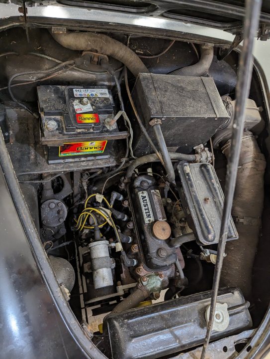 1958 Austin A35 - the family heirloom... - Page 1 - Readers' Cars - PistonHeads UK - The image shows a close-up of the engine bay of a car, featuring various mechanical components. There is a battery in the center with a yellow and red label on it. To the right of the battery, there is a black box with an orange sticker on it, labeled "Battery". Below this box, there are several wires and connectors, including what appears to be a starter motor and various cables connecting it to other parts of the vehicle. The engine bay is dirty and rusty, suggesting that the car may be old or not well-maintained. The image captures a detailed view of the car's internal mechanics, with visible gaskets and other automotive components.