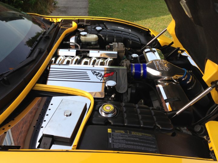 2001 C5 Targa Top value - Page 1 - Corvettes - PistonHeads - The image showcases the open hood of a bright yellow car, revealing the intricate workings beneath the hood. The engine is pristine and well-maintained, with various metal parts, black battery, and blue air filter clearly visible. There's a noticeable accessory on the battery, possibly for jump-starting. The car is parked on what appears to be a residential street, with a green lawn filling the background.