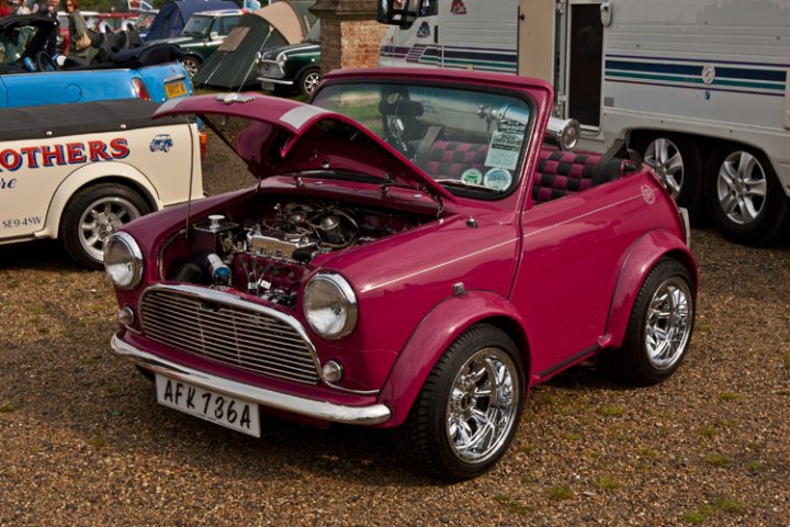 London to Brighton  - Page 1 - Classic Minis - PistonHeads - The image showcases a classic Mini Cooper car that has been customized with a lot of chrome. The car is parked on a gravel surface with a dirt ground visible, and its hood is propped open, revealing an intricate engine with many chrome details. In the background, there are other vehicles, including an RV and other classic cars, suggesting this could be a car show or a gathering of vehicle enthusiasts. The red and grey color of the car adds to its vintage charm.