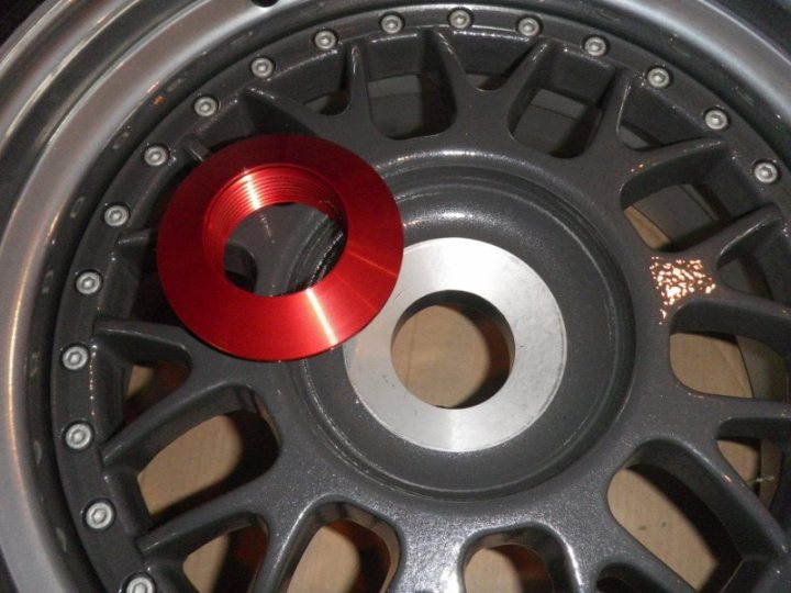 Seized wheel nut and an unhappy gear display - Page 1 - Radical - PistonHeads - The image presents a close-up view of a hubcap, focusing on its central hub. Prominently, a large, bright-red hole is encircled by a series of bolts. The hubcap itself appears to be made of metal, with a shiny, polished surface that reflects light. The overall color tone of the image is somewhat metallic, with the red hole standing out against the more neutral colors of the hubcap and the background.