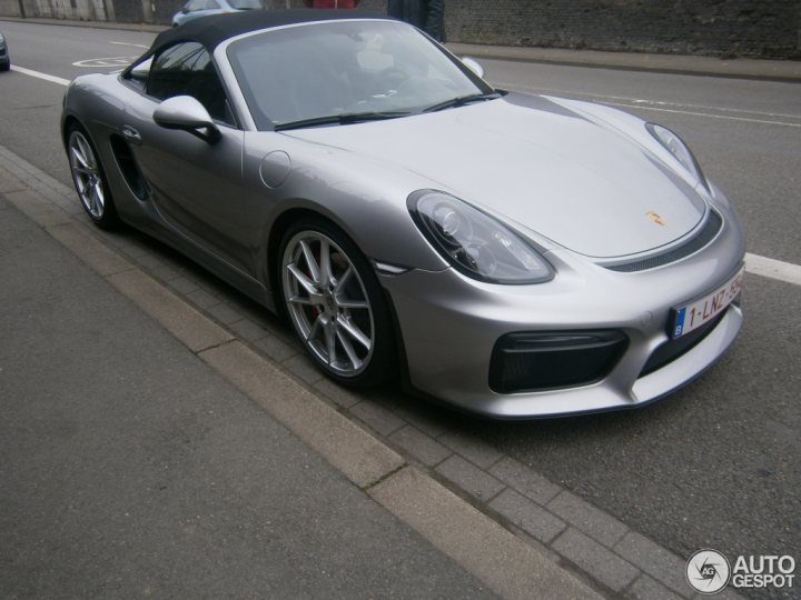 A black and white photo of a car parked in a parking lot - The image captures a scene on a street where a silver Porsche sports car is the main focus. The car's sleek design and shiny metallic surface reflect the light, suggesting it could be during the day although the exact time is not clear. The Porsche is parked on a curb next to a sidewalk, adjacent to a designated parking zone. A single red license plate is visible on the car, adding a pop of color to its otherwise monochromatic exterior.