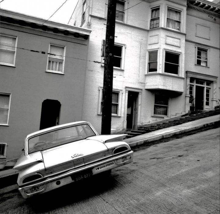 A 'period' classics pictures thread (Mk III) - Page 33 - Classic Cars and Yesterday's Heroes - PistonHeads UK - The image showcases a vintage scene featuring an old car parked in front of a two-story building. The architecture of the building suggests it might be an older structure with traditional design elements, such as a gable roof and vertical siding. The car has a classic design, reminiscent of models from the mid to late 20th century, and appears to be in good condition. The street is lined with residential houses that share a similar architectural style, creating a cohesive neighborhood aesthetic. There are no visible people or signs of movement, giving the scene a quiet and still atmosphere. The overall impression is one of a bygone era captured in this snapshot of urban life.