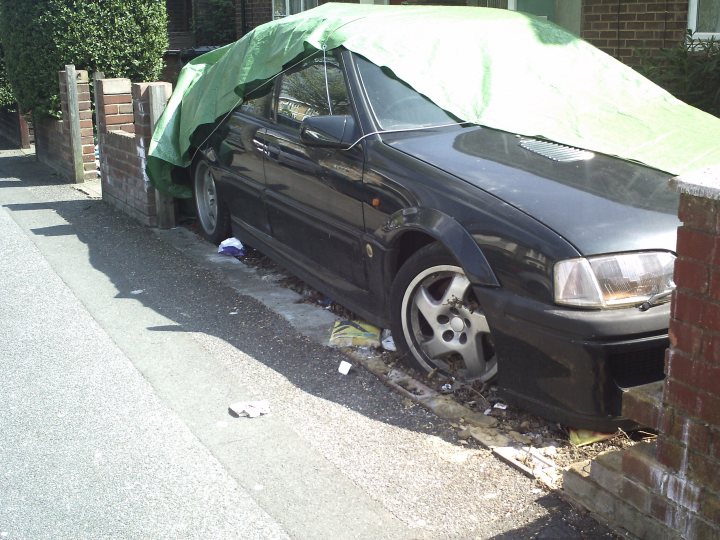 Pistonheads Side Road Relics - The image depicts a dark-colored car parked on the side of a street. The car is partially covered with a green cover, possibly a flexible fabric or a canvas tarp. This suggests that the car might be hidden from view, or might be protected from the elements. Some debris and trash are scattered around the pavement under the car, implying that the street might not be well-maintained. There is also a brick wall to the right of the car, which adds to the urban setting of the scene. The sunlight casts shadows both on the ground and on the car, suggesting that the photo was taken during the day with clear skies.