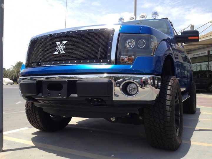 A white truck parked in a parking lot - Pistonheads - The image shows a large blue pickup truck parked in a lot. It features a chrome grille with a letter X in the center, which is a well-known and stylish design choice for the make and model of the X lineup. The truck has a black radiator grate and its chrome detailing on the hood and along its side strikingly contrasts with the blue paint. The tires are large, presumably for its utility purposes, and the big, round headlights suggest off-road capabilities. The time is bright daylight, without any immediate notice of clouds or rain, which gives the photo a clear and vibrant feel.