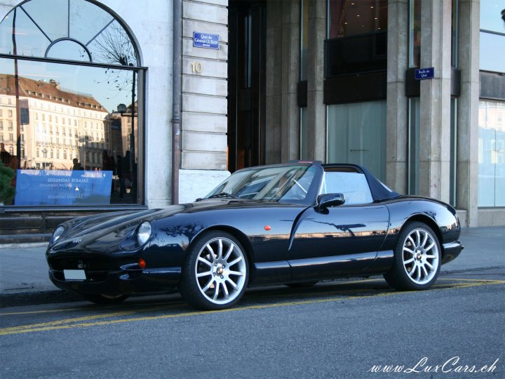 Pics of Chimaeras with after-market alloys - Page 17 - Chimaera - PistonHeads - The image shows a black sports car, the Maserati 320, parked on a sidewalk in front of an old city building. The car is positioned perpendicular to the curb, and its sleek design and silver rims give it an elegant and expensive appearance. The building behind the car is ornate, with large windows reflecting the nearby trees and a street sign. On the sidewalk, there's some debris which appears to be litter. A blue "M" can be seen on the building, possibly indicating the renovation area.