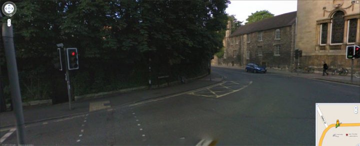 Pistonheads - The image portrays a quiet, narrow street with buildings on both sides. A few pedestrians are visible, going about their day. The street is lined with lush greenery, providing a serene atmosphere. Various traffic lights are present at various points down the street, adding to the urban feel despite the calm setting.