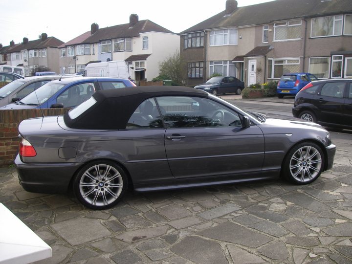 Pistonheads - The image shows a two-door black car parked on a slate-gray paved area in front of residential housing. The car has a soft top roof and aftermarket spoilers. In the background, there's a clear sky and other cars parked along the street. There is potted greenery near the car, and the overall setting suggests a quiet residential area.