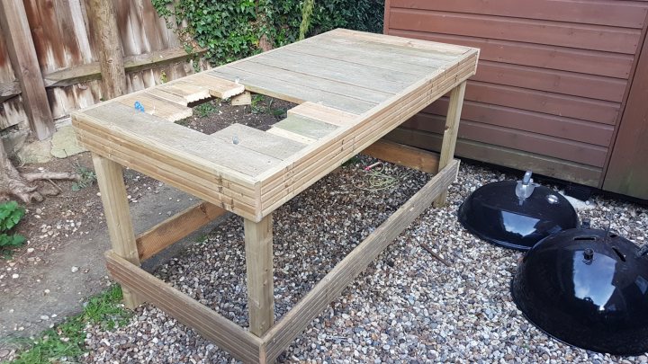 Pallet wood Weber grill table project on a budget - Page 1 - Homes, Gardens and DIY - PistonHeads - The image shows a wooden garden bench or picnic table. The table is unfinished and appears to be placed on a gravel surface. There is greenery in the background, suggesting the setting is outdoors. There are two objects that resemble fire hydrants near the table, although their exact nature is not clear. The table has a flat surface with visible splits, indicative of it being a piece of second-hand furniture.