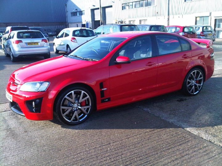 My New Car....... - Page 1 - East Anglia - PistonHeads - The image is a photograph of a red coupe parked in a parking lot. The vehicle is sleek and modern in design, featuring alloy wheels and a sporty body shape. It is angled to show its front left side, with the sun reflecting on its shiny paint. The background reveals a commercial area with industrial buildings and other parked cars. There is a license plate visible on the front of the car. The style of the photo appears to be a casual, unprofessional shot, capturing the car from a slightly elevated angle to accommodate the height of the vehicle.