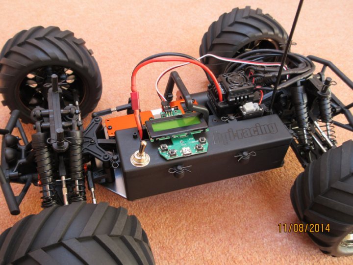 The 1/10 offroad RC thread - Page 1 - Scale Models - PistonHeads - The image shows a remote-controlled vehicle with a prominent, square-shaped battery module. The battery module appears to have electronic components and a screen displaying "EIGHTNET" and "XYZVehicle," suggesting it's a part of an electronic system like radio-controlled cars or boats. The vehicle is resting on a carpeted floor with a brownish hue. The rotating wheels, textured like tires, indicate this is not a flat or smooth surface typical for indoor environments. There is a red wire connected to the battery module, possibly for charging or power supply.