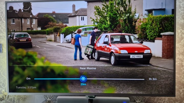 Incorrect number plates in TV and Film - Page 3 - TV, Film, Video Streaming & Radio - PistonHeads UK - This image shows a living room scene where a large flat screen TV is displaying a scene from a television show. The TV, which occupies the upper right corner of the image, has the title "The Walk" written at the bottom and indicates that it's playing Episode 1.

On the TV screen, we can see two individuals standing in front of a car parked on the side of a street. One person is holding a handbag while the other person appears to be engaged in conversation with them. The car they are near has a red and white color scheme, suggesting it might be an older model vehicle.

In the foreground of the image, there's a glimpse of a living room setting, with a small portion of furniture visible, including what seems to be a sofa or chair in the lower left corner. The overall ambiance suggests a casual, comfortable home environment.