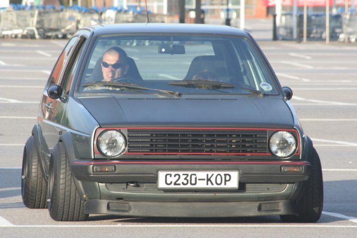 Pistonheads Scene Dub - The image captures a scene of an old black Volkswagen beatle driving across a parking lot. The Volkswagen, characterized by its iconic rounded shape and small size, has a distinctive red stripe on its side. Its license plate, reading "C230-KOP," is clearly visible. Inside the vehicle, there are two individuals, one driving and another seated in the passenger seat. Both are wearing sunglasses, suggesting either the sun is out or it's just their style. The license plate and the stylish sunglasses worn by the occupants add a distinctive and personal touch to this everyday scene.