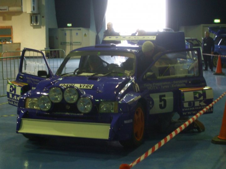 Pistonheads Weekend Stages - The image depicts an indoor scene, likely a garage or warehouse, with a prominent vintage blue Volkswagen Beetle in the foreground, characterized by its distinctive rounded shape, large headlights, a series of round headlights on the front bumper, and number "5" written on its side. Behind the Beetle, there is an orange security cone. Further to the right, there is a person standing in the background behind a network of barricades or netting. Additional blue vehicles and exposed boxes are visible in the distance, indicating a potentially large shared space or exhibition area. The overall atmosphere suggests a casual or open-to-the-public event.